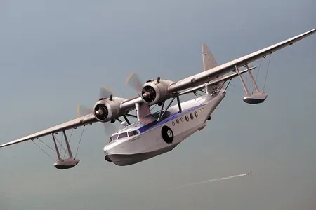 Silver twin engine flying boat over a vast ocean. A small boat leaves it's wake below.