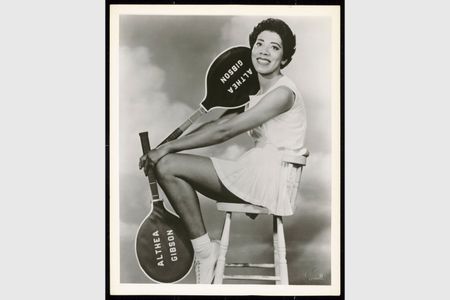 Althea Gibson poses on a stool with two tennis rackets. Both the rackets have dark colors printed with "Althea Gibson" in white letters.