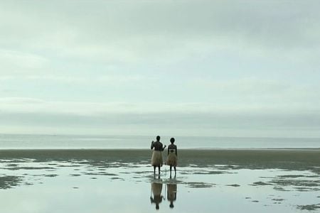 Two people in grass skirts stand on the shore, facing the ocean under an overcast sky.