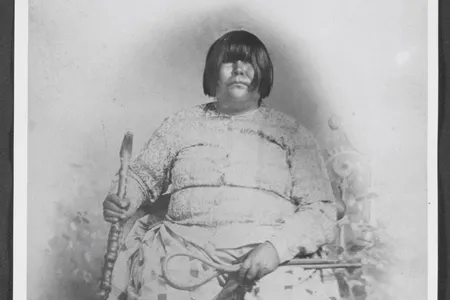 Black and white portrait of Dat So La Lee seated in a chair and surrounded by six decorative baskets on the floor by her feet.
